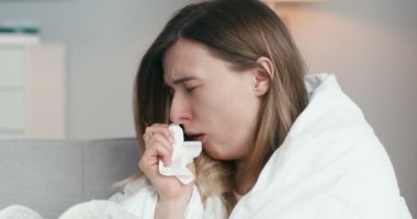 Hasta, kötü görünümlü, beyaz bir kadının evinde, kanepede oturup, hapşırması, hadım etmesi ve peçeteye burnunu silmesi. Coronavirus konsepti. Grip belirtileri gösteren bir kadın..