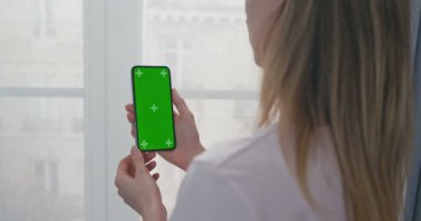 Dikey akıllı telefonlu, yeşil ekranlı bir kadının omzunun üstünden bak. Kendini izole etme penceresinde duran, video izleyen ve telefonda sohbet eden bir kız. Videochat. Krom anahtar. İzleme hareketi.