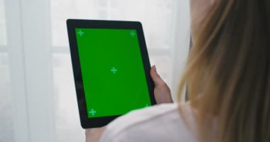 Dikey tablet bilgisayarlı, yeşil ekranlı bir kadının omzunun üstünden bakın. Kendini izole etme penceresinde duran bir kız, video izleyip, web kamerasıyla görüntülü sohbet ediyor. Videochat. Krom anahtar.