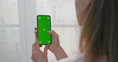 Siyah, dikey, yeşil ekranlı akıllı telefondan kadının omzunun üstünden bak. Pencerenin önünde duran ve cep telefonunun haber sayfasını tıklatan ya da kaydıran kız. Kapatın. İzleme hareketi. Kapalı.