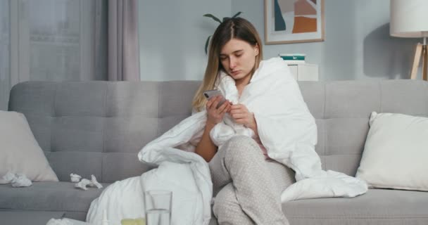 白种人年轻的生病妇女穿着毛毯坐在客厅的沙发上 蜷缩在餐巾纸上 在手机上拍拍或滚动新闻 验尸官的概念 感染流感的女孩在智能手机上发短信 — 图库视频影像
