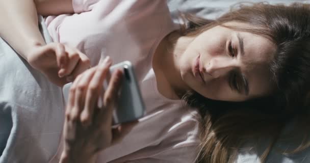 一个年轻的白人美女躺在床上 躺在毯子下 用智能手机聊天 发短信 在卧室里看美女在手机上窃听器和滚动的头像 从上面看 — 图库视频影像