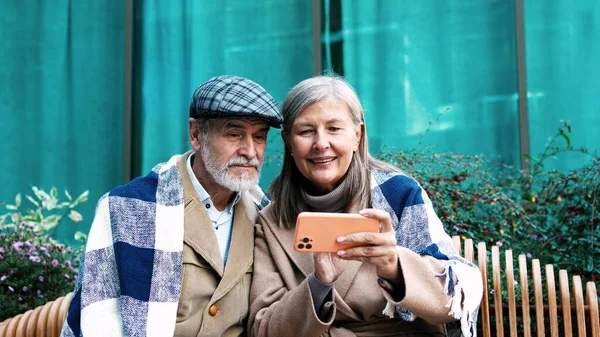 Lovely happy married couple of elderly handsome grey-haired man sitting on bench outside with beautiful cute mature woman wearing coat, plaid video calling using smartphone. Communication concept.