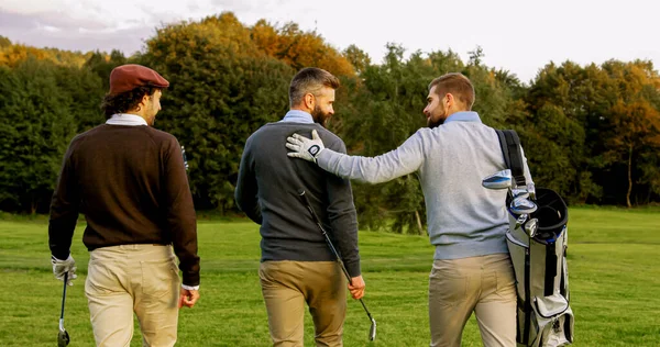 Three male friends with clubs and golf bag walking on the pitch. Back view