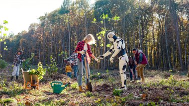 Close up of robot humanoid helping to human in planting seedlings of trees in field at forest or park. Sunny fall day. Android interacting with woman for saving forests and eco planet. Outdoor.