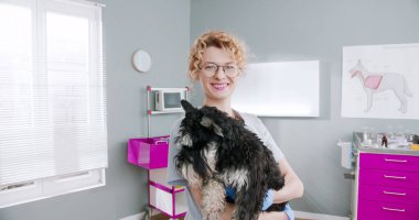 Close up of female veterinarian with gloves holding cute dog and looking into camera. Vet standing in medical suit with stethoscope. Concept pets care, veterinary