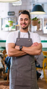 Vertical video of cheerful young Caucasian man worker in apron standing in restaurant, looking at camera and smiling at work. Male barista in coffee shop with smile on face. Small business owner