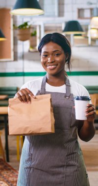 Portrait of happy beautiful young African American woman barista smiling while standing in restaurant looking at camera holding take away order and coffee-to-go in hands. Vertical video small business