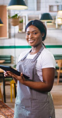 Portrait of African American young joyful beautiful woman worker in apron surfing internet typing on tablet device standing in coffee shop and smiling looking at camera. Vertical orientation photo