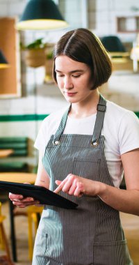 Portrait of Caucasian young joyful beautiful woman waitress in apron surfing internet typing on tablet device standing in coffee shop and smiling looking at camera.