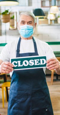 Portrait of old nice grey-haired Caucasian male worker in apron wearing medical mask looking at camera standing in coffee house holding in hands Closed sign business in lockdown Vertical video concept