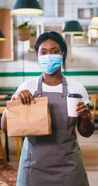 Portrait of young beautiful African American woman employee in medical mask and apron standing in coffee house looking at camera holding package with take away order and coffee-to-go Vertical footage
