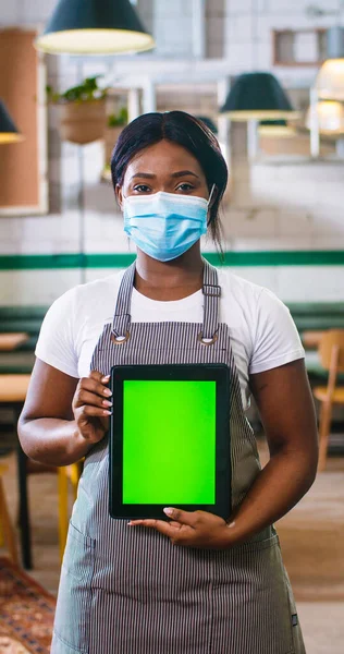 Vertical orientation of happy young African American beautiful woman worker in medical mask standing in cafe at workplace in apron, looking at camera, holding tablet with green screen, chroma key