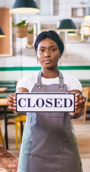 Vertical photo of African American young beautiful female employee wearing apron standing in restaurant holding in hands Closed sign, waitress at work in restaurant, closing small business, portrait