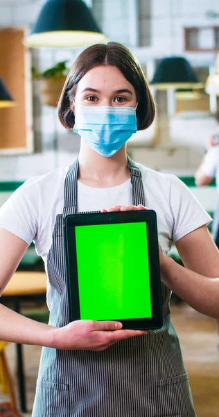 Vertical orientation photo of happy young Caucasian beautiful woman worker in medical mask standing in cafe at workplace in apron, looking at camera, holding tablet with green screen, tech concept