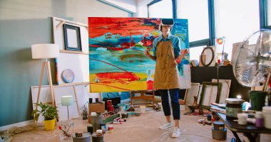 Futuristic Caucasian female painter in apron in studio wearing virtual reality headset creating masterpiece. Modern artwork paint on canvas, contemporary and successful fine art artist in VR glasses