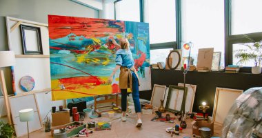 Rear of innovative Caucasian young female painter drawing with her hands on big canvas, using fingers. Contemporary artist creating colorful, sensual oil painting artwork in art studio, art concept