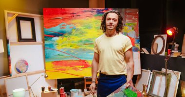 Caucasian young handsome contemporary man holding palette with paints turning and looking to camera in own art studio at night. Workshop concept