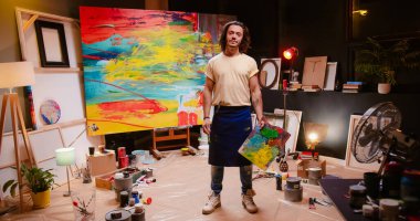 Caucasian young handsome contemporary man painter looking at finished colorful painting holding palette with paints turning to camera in own art studio at night. Workshop concept