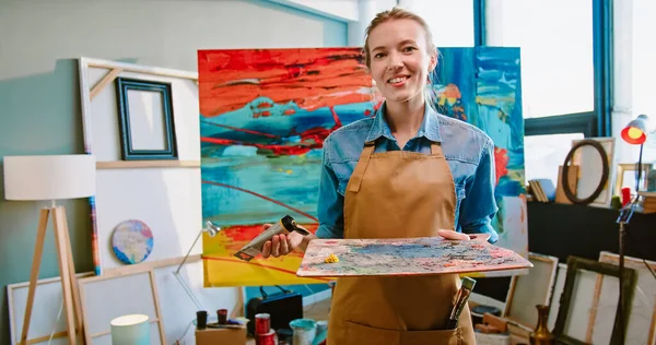 Portrait of cheerful Caucasian young pretty happy woman artist stands in workshop alone smiling and pouring paint on palette before painting on canva. Modern artwork, painter at work, hobby concept