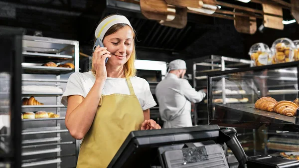 Caucasian female seller in bakeshop standing at paydesk and talking on mobile phone. Beautiful woman baker in apron and hat speaking on cellphone while selling bakery. Workday. Telephone call.