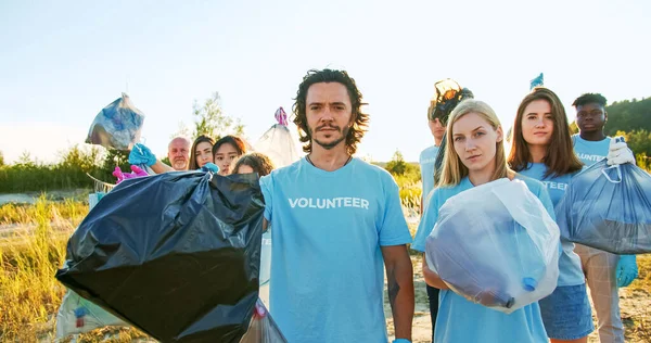 A group of nature activists after garbage collection work. Members of a voluntary organisation looking at the camera and lifting rubbish, trash bags. Nature, ecology protection concept.