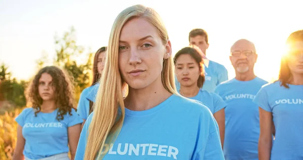 Young attractive woman with a group of eco activists in the background. Pretty Caucasian female volunteer looking at camera and saying: I Am a Volunteer. Volunteering, environmental conservation.