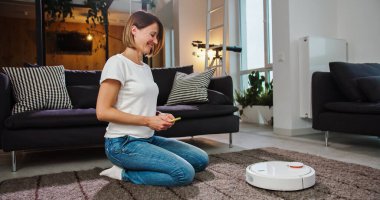 Neşeli genç bayan akıllı telefon kullanarak robotik süpürgeyi açıyor ve otomatik temizliğin tadını çıkarıyor. Modern teknoloji ve gençlik, temizlik, temizlik, yaşam tarzı konsepti.