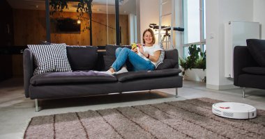 Beautiful young lady is turning on vacuum and enjoying automatic cleaning sitting on sofa and relaxing with smartphone. Modern technology and youth concept.