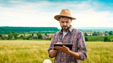 Beyaz şapkalı yakışıklı adam çayırda duruyor ve tablet kullanıyor. Açık havada. Yakışıklı genç çiftçi elinde bilgisayarla tıkırdıyor ve kaydırıyor. Çiftçilik için araç.