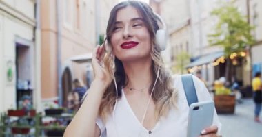 Kulaklıkla müzik dinleyen çekici genç bir kadının portresi. Şehir yürüyüşünde akıllı telefon kullanarak etrafa bakıp gülümsüyor. Güzel bir gülümseme. Güzel gözler ve kırmızı ruj..