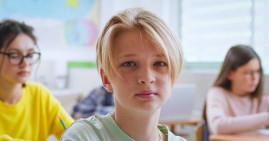 Sınıf arkadaşları arka planda masa başında otururken kameraya bakan güzel bir çocuğun portresi. Modern okulda ders sırasında dikkatle dinleyen zeki çocuklar..
