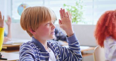 Gündelik giyinen sevimli çocuk, yükselen el kaldırıyor ve öğretmenlerin sorularını cevaplıyor. Akıllı genç erkek öğrenci iyi notlar almak istiyor..