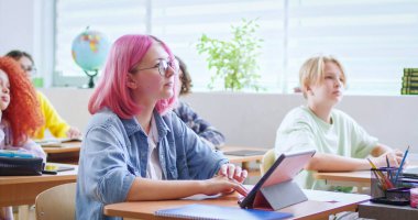 Gözlüklü, pembe saçlı, tablet cihazıyla masada oturan ve kameraya gülümseyen mutlu beyaz kız portresi. Okulda bilgisayar kullanan neşeli stil sahibi bayan öğrenci. Ders..