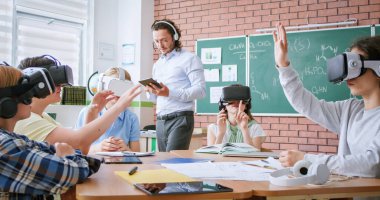VR kulaklıklı bir grup genç öğrenci masada otururken dijital tabletli erkek öğretmenler eğitim programını açıklıyorlar. Okulda etkileşimli ders sırasında modern teknolojiler.