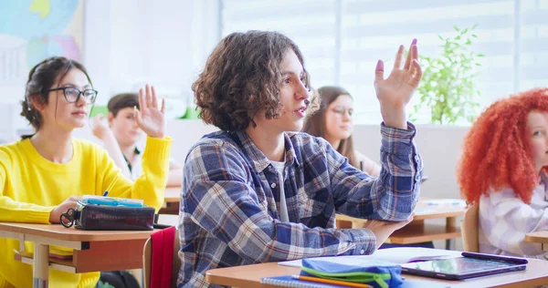 积极的高加索男孩 波浪形的黑发举起手 在课堂上回答问题 有信心的男生与背景的同学坐在课桌前 — 图库照片