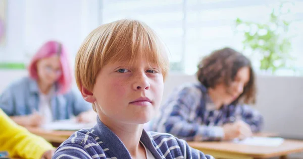 一个漂亮的白人聪明的小男生坐在教室的书桌前 看着镜头看课时的肖像 在孩子们中间室内 小男孩在学习学校的概念 靠近点 — 图库照片