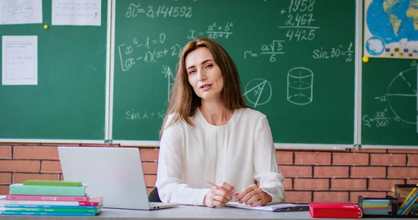 魅力的な女性数学教師は 現代の教室でラップトップで机に座っています カメラを見ながら三角形の紙シートを持って話す自信のある女性 — ストック写真