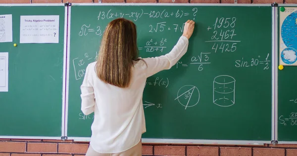 Back view of female teacher standing near green chalkboard and writing geometric formula in classroom. School tutor solving and explaining mathematics rules.