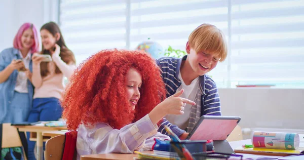 stock image Side view of boys and red haired girl talking and using digital tablet during break in school. Happy classmates in casual attire discussing funny video in bright classroom.