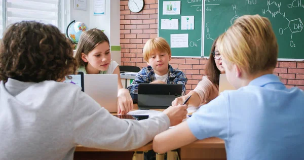 stock image Enthusiastic female teacher using laptop for explaining chemical compounds to children during lesson. Attentive pupils looking at task on sheet of paper and making notes in workbook.