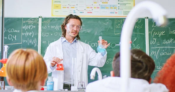 Caucasian man in glasses and lab coat holding test tubes with colored liquids and explaining pupils chemical reactions. Education with interest and modern school concept.