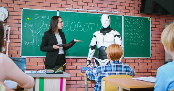 Caucasian female teacher with tablet in hands presenting humanoid robot to attentive pupils at classroom. Interactive lesson at modern school.