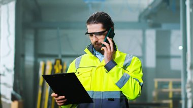 Gözlüklü ve sarı üniformalı beyaz erkek mühendis fabrikada duruyor ve cep telefonuyla konuşuyor. Mühendislik konsepti. İçeri gir. Fabrikadaki dosya klasörüyle cep telefonundan konuşan adam..