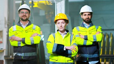 Kasklı ve üniformalı, kameraya gülümseyen ve el ele tutuşan beyaz erkek ve kadın meslektaşlarının portresi. Üretim alanındaki erkek ve kadın iş arkadaşları. Fabrika işçileri takımı.