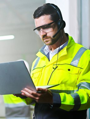 Kafkasyalı, kulaklıklı ve üniformalı bir adamın dikey görüntüsü. Manufactory 'da dikey görüntüsü, video kaset ile konuşuyor ve dizüstü bilgisayarda çalışıyor. Mühendislik konsepti. Bitki videosundaki adam bilgisayarda sohbet ediyor..