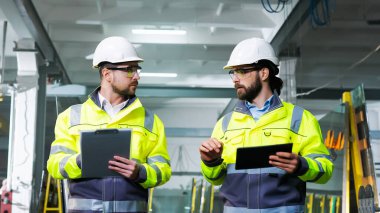 Erkek iş arkadaşları üretim fabrikasında yürürken konuşur ve plan yapar. Üretimdeki belgeler ve tablet cihazları klasörüyle çalış. Üretim ortak çalışma. Uzmanların tartışması.