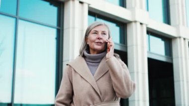 Telefonda konuşan mutlu, gri saçlı çekici bir kadının portresi. Başarılı emekli, ceketli, dışarıda yürüyen, akıllı telefondan gülümseyen bir kadın..