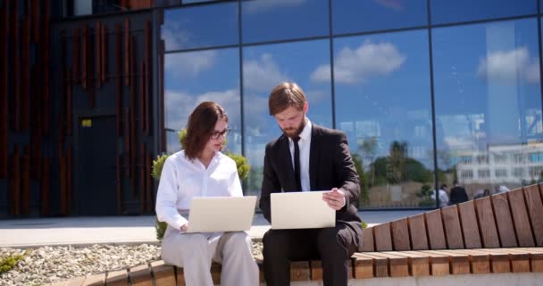 高加索商人和女商人坐在现代办公大楼附近的街上 一边谈论和讨论工作 一边在笔记本电脑上工作 讨论工作的男人和女人 — 图库视频影像