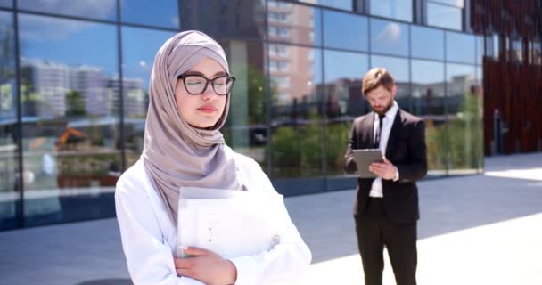 ヒジャーブのアラビア人イスラム教徒女性の肖像画と 現代のオフィスビルでカメラに笑顔のメガネ バックグラウンドでタブレットデバイスを持つ白人ビジネスマン 平等な権利 平等か アラブ ビジネスウーマン — ストック動画